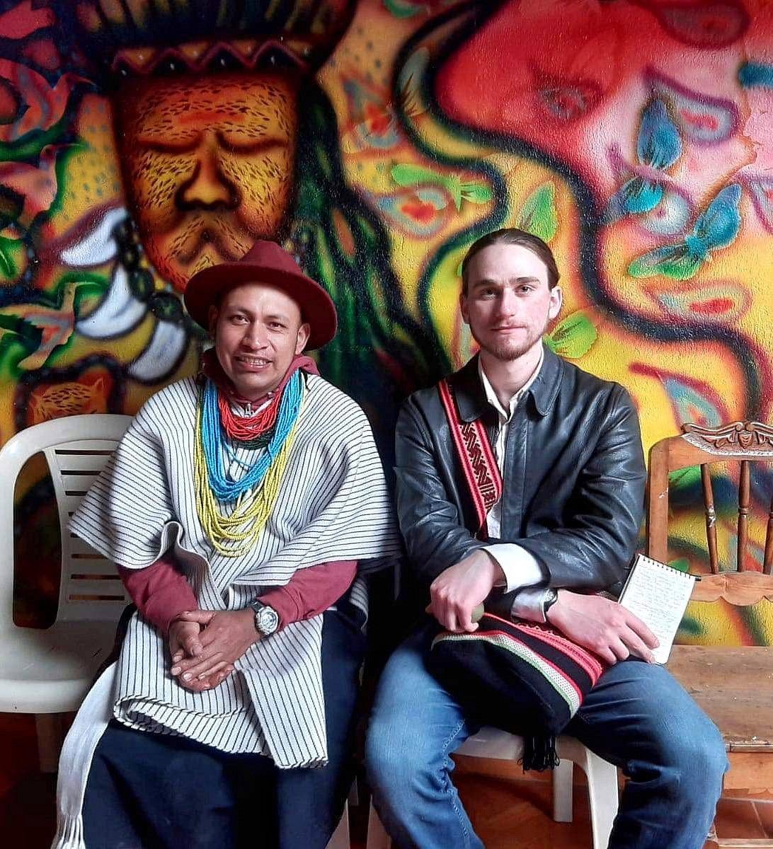 Rowan Glass seated with shaman friend with a colorful mural in the background 