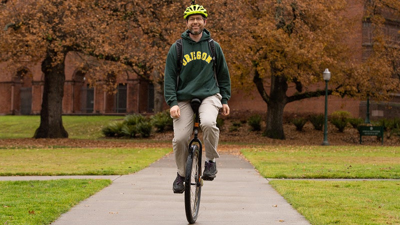 Danield Lowd, in UO hoodie on campus, rides a unicycle toward camera