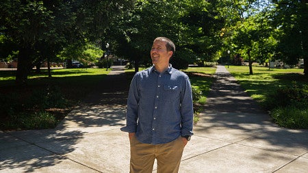 Raoul Liévanos walking on tree-lined campus, smiling amid a sunny day