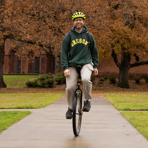 Danield Lowd, in UO hoodie on campus, rides a unicycle toward camera